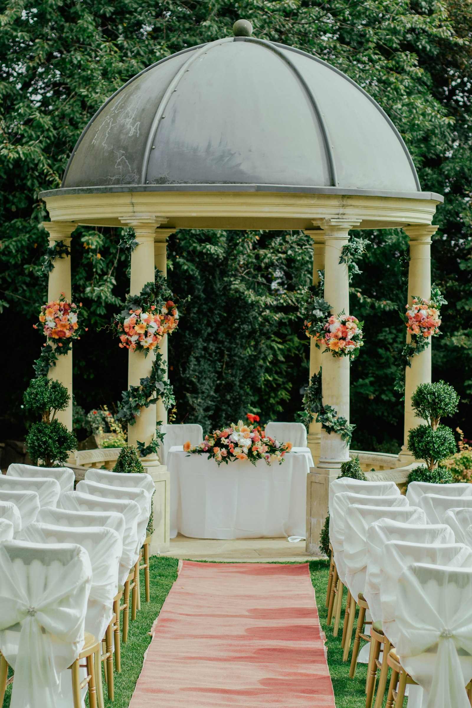 A garden ceremony with a domed stone gazebo, flowers and rows of white chairs
