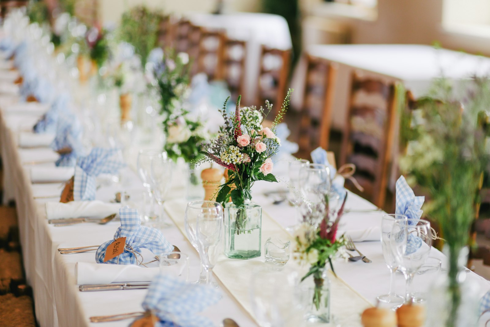 A long reception table set with white linen, blue gingham napkins and wildflowers